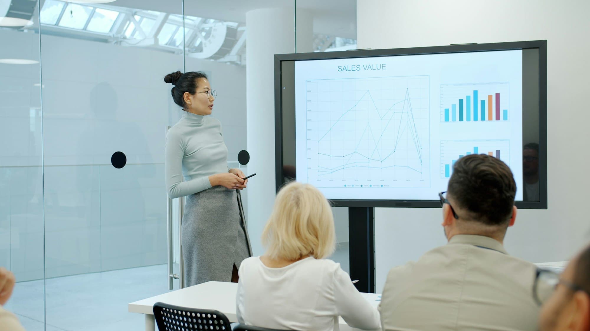 A speaker presenting a chart on a large screen to two colleagues in a bright glass meeting room.