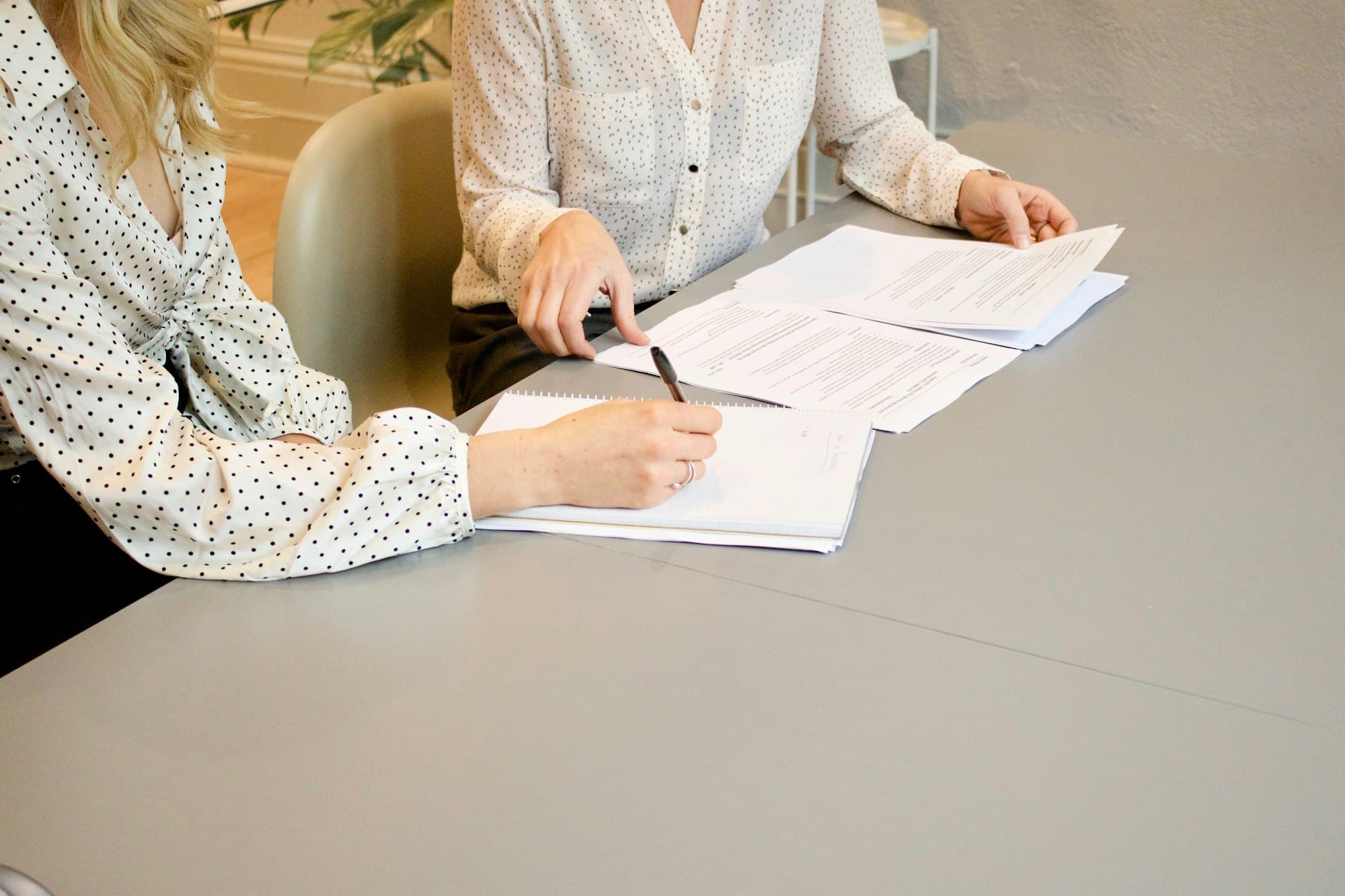 Two people at a light grey table reading printed pages together, one marking a sheet with a pen.