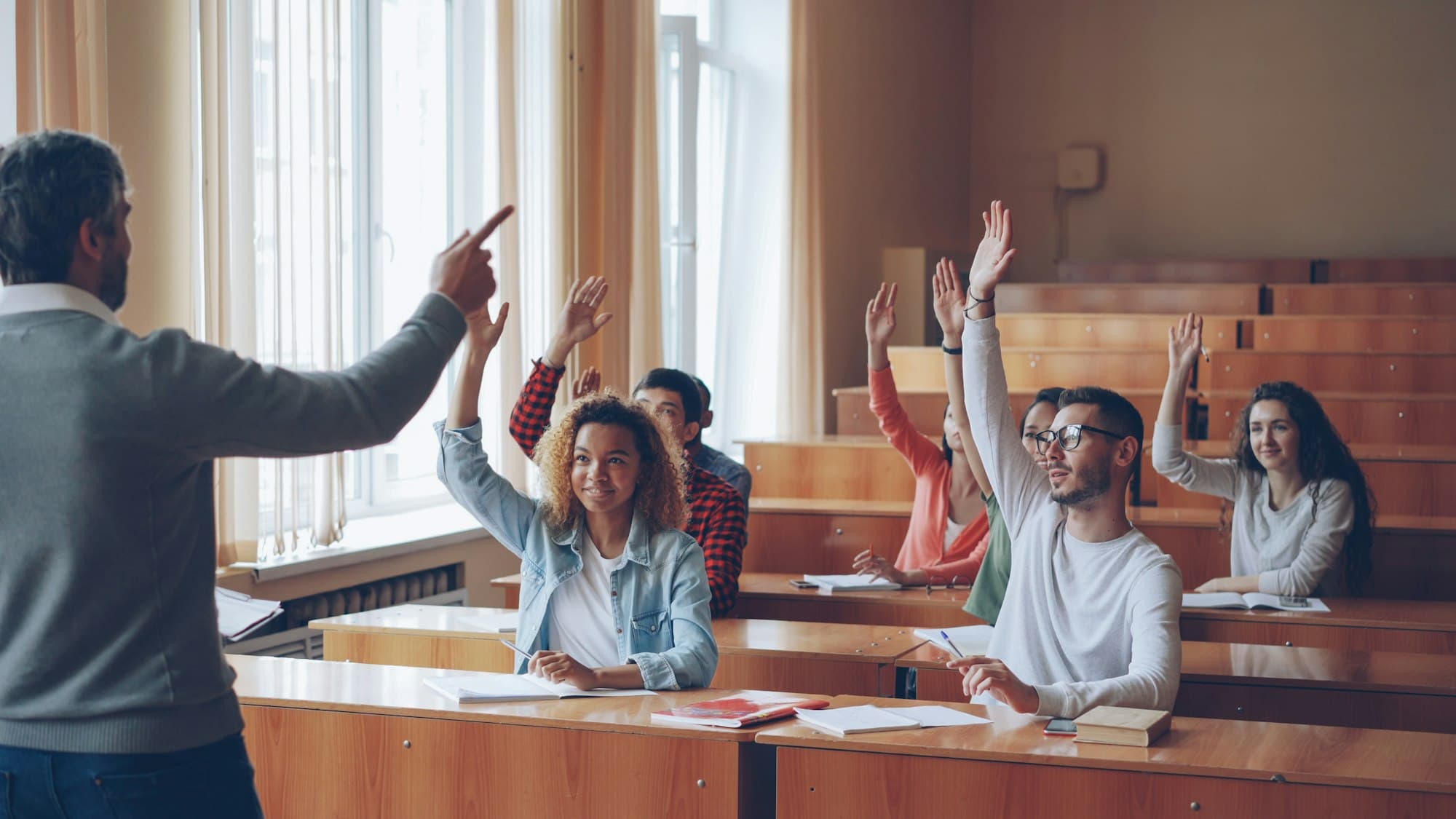 Students raising their hands in a lecture hall as a lecturer points to one of them.